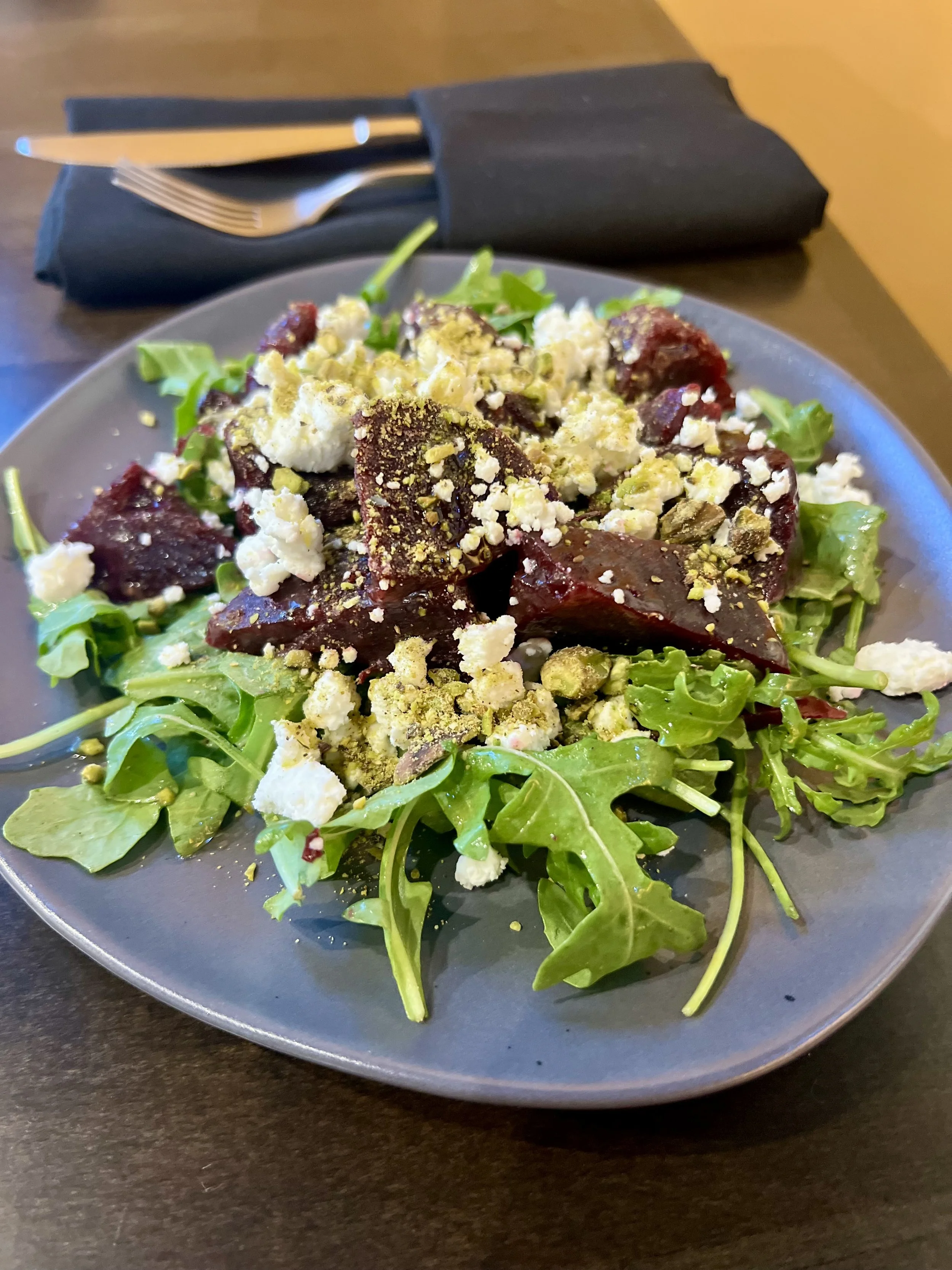 Salad with leafy greens, roasted beets, crumbled feta cheese, and crushed pistachios on a gray plate with black napkin and cutlery in the background.