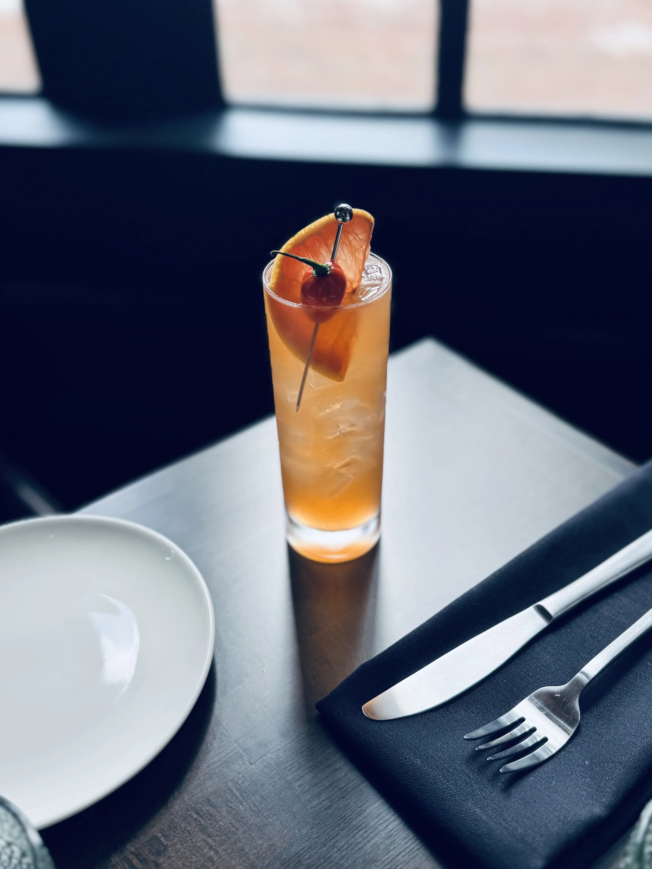 A tall glass of orange cocktail garnished with an orange wedge and cherry, set on a table with a white plate, a black napkin, a knife, and a fork, near a window with a blurry view outside.