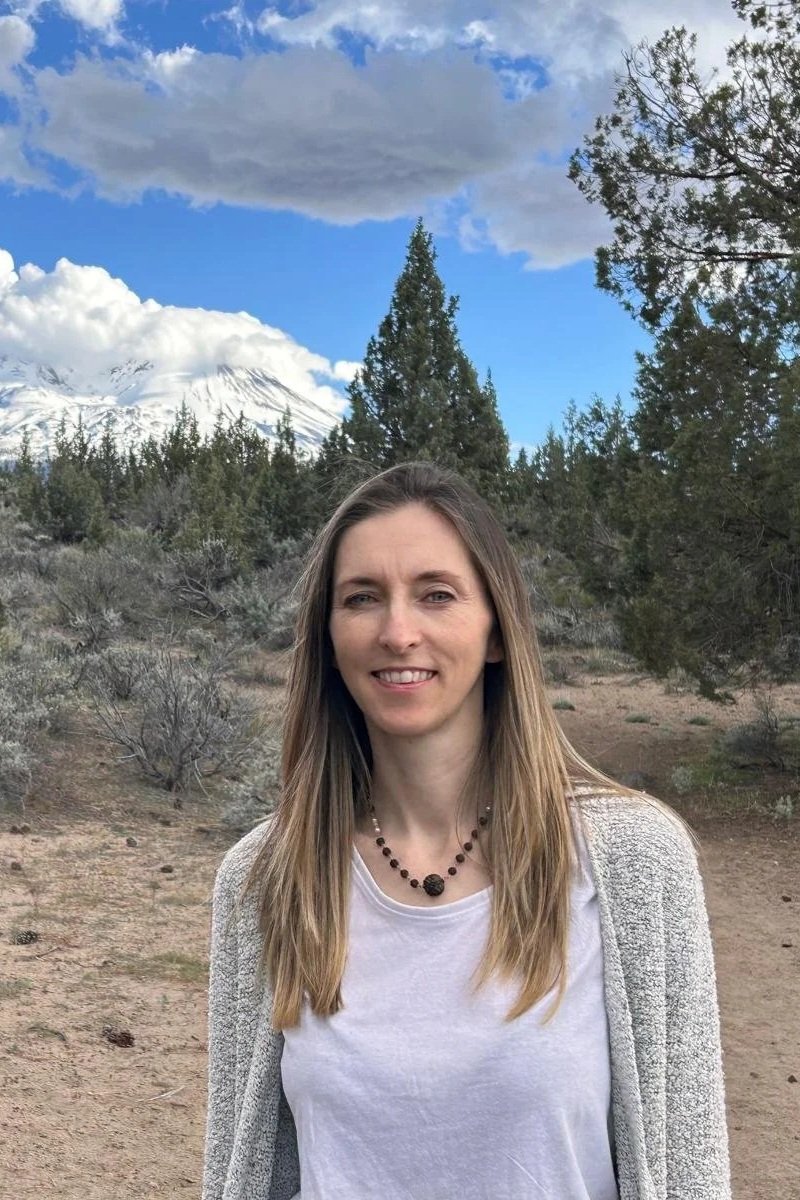 A woman with long, light brown hair, wearing a white shirt and gray sweater, standing outdoors in a desert-like landscape with trees and shrubs, snow-capped mountains in the background, and a partly cloudy sky.
