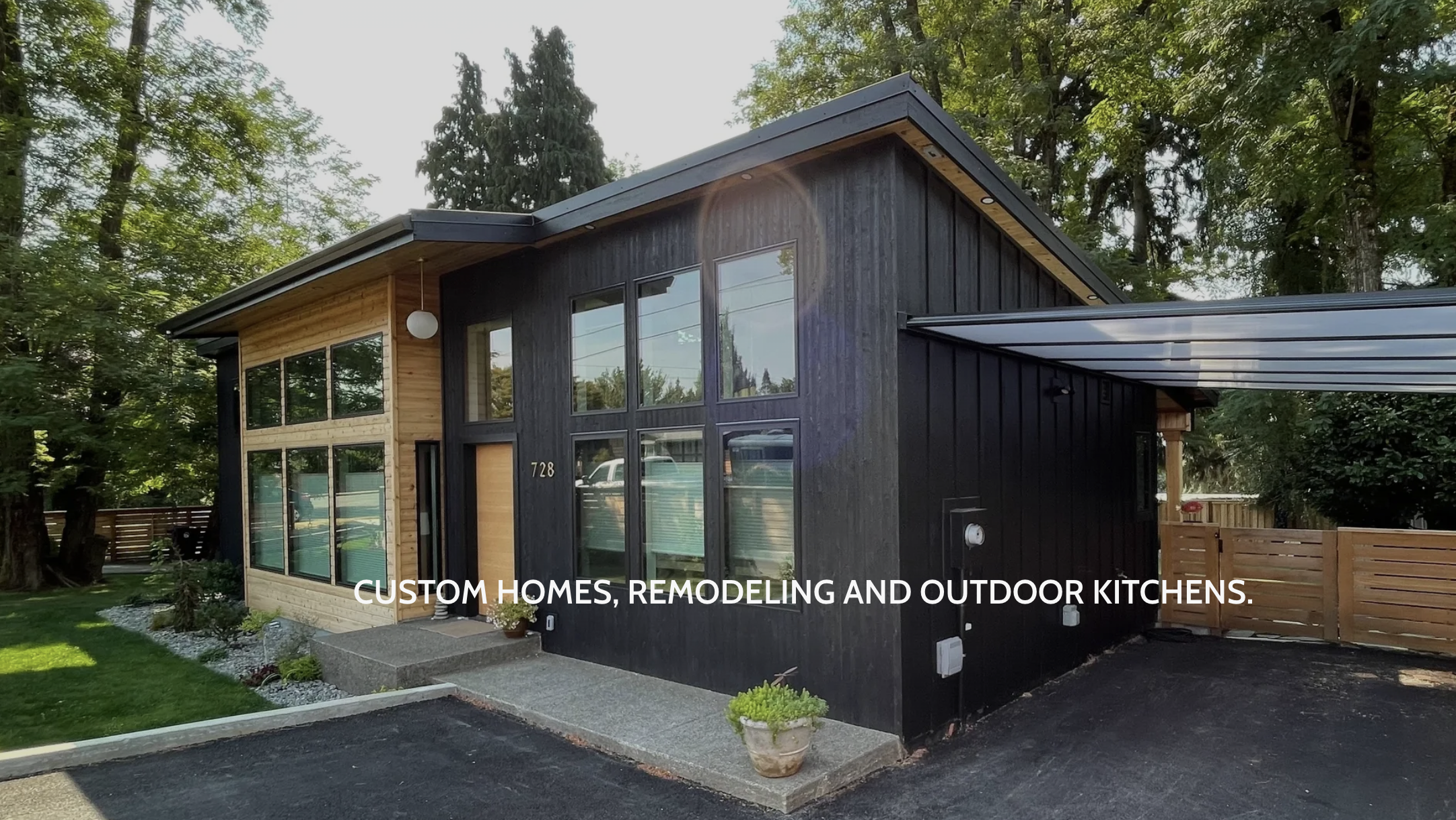Modern two-tone house with large windows, a driveway, and a small garden with shrubs and potted plants. The house has black and wooden exterior siding, surrounded by tall trees.