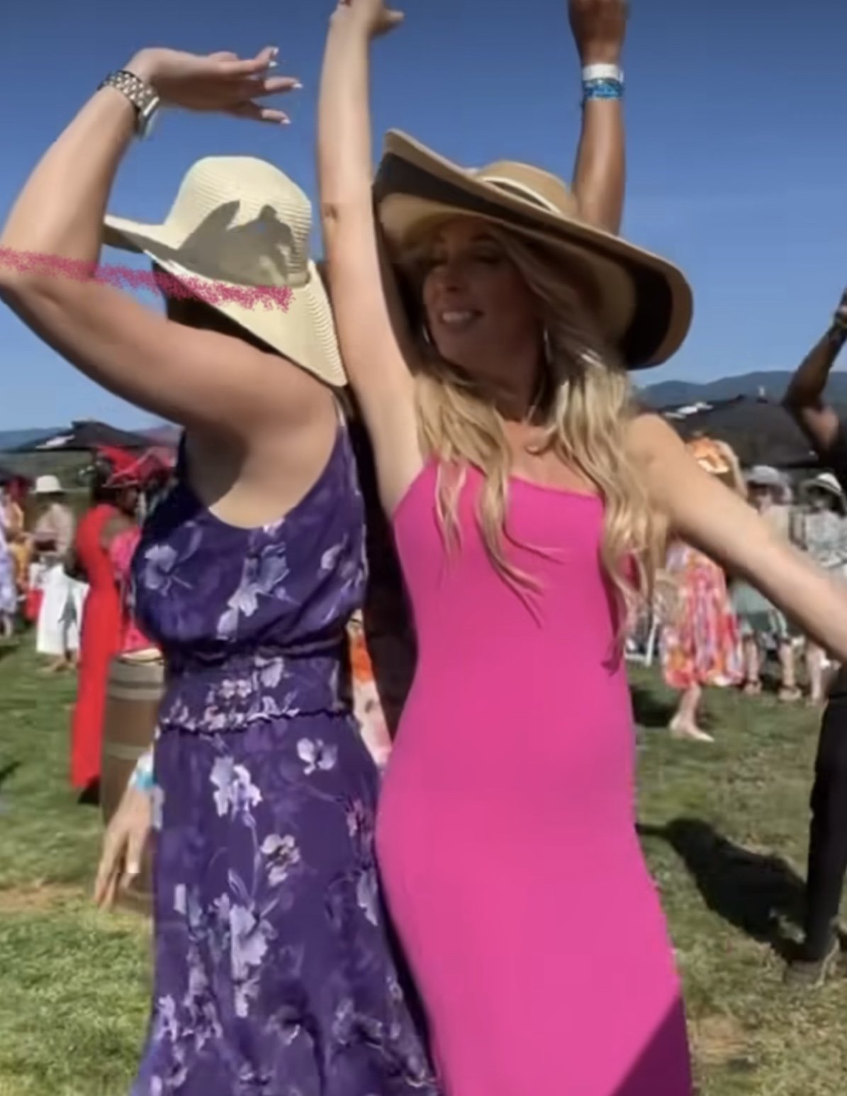 Kelly Craig Graham and friends enjoying a Polo party in Seattle Washington.  dancing outdoors on a sunny day, both wearing wide-brimmed hats, one in a purple floral dress and the other in a pink dress, with a crowd in the background.