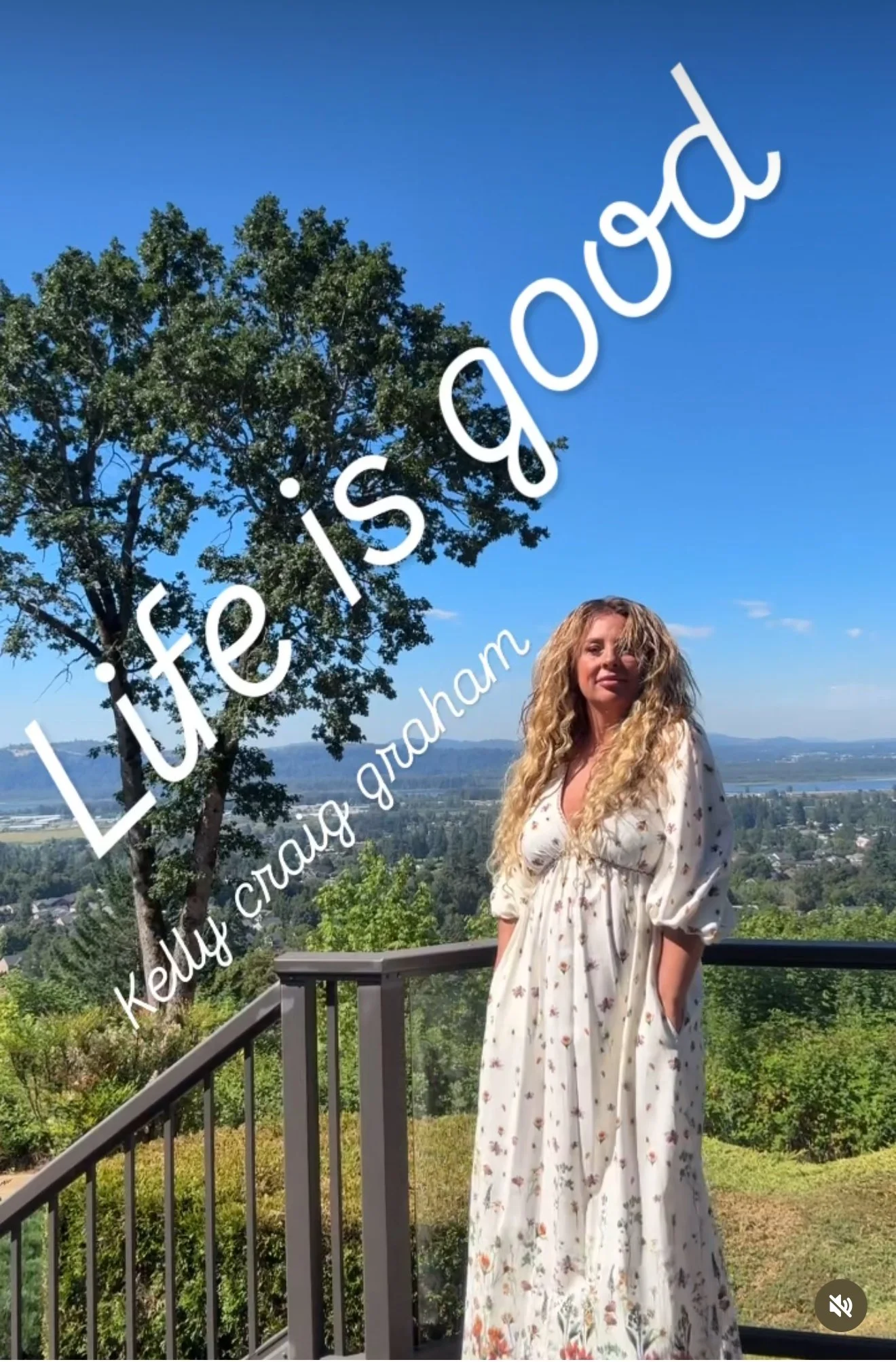 A woman with curly blonde hair standing on a balcony overlooking a lush landscape with trees, hills, and water under a clear blue sky. Overlaid text reads, 'Life is good' and 'Kelly Craig Graham.'