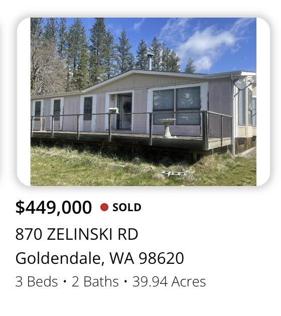 Exterior view of a single-story house on a grassy lot with trees in the background, enclosed by a wooden deck and railing.