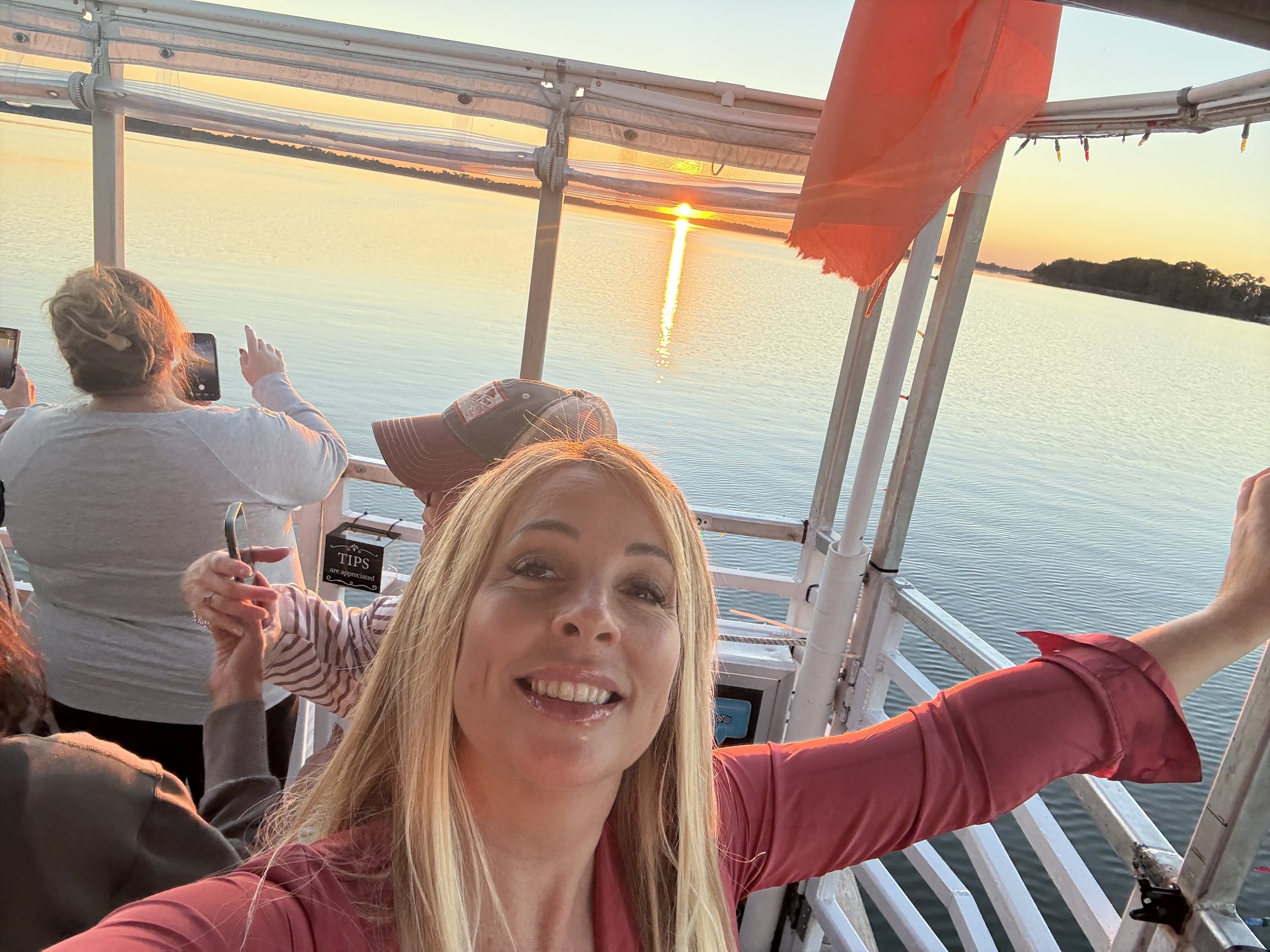 Kelly Craig Graham smiling and taking a selfie on a boat at sunset, with other passengers in the background, over a body of water with trees on the far shore. Enjoying time with friends in Florida.