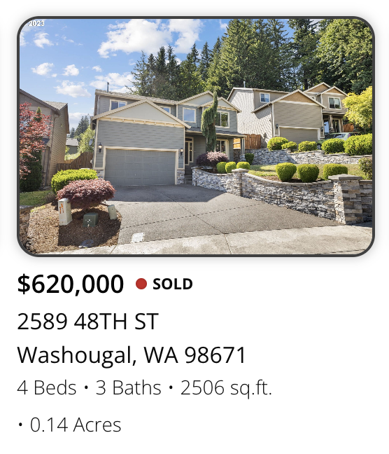 Photo of a multi-story house with a gray garage door, landscaped front yard with bushes and trees, and a stone retaining wall. The house is surrounded by tall trees and a partly cloudy sky.