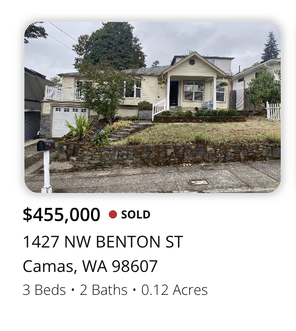 Front view of a house on a hill with a stone retaining wall, stairs, and a small front yard with bushes and a tree. The house is white with a porch and a garage to the left.