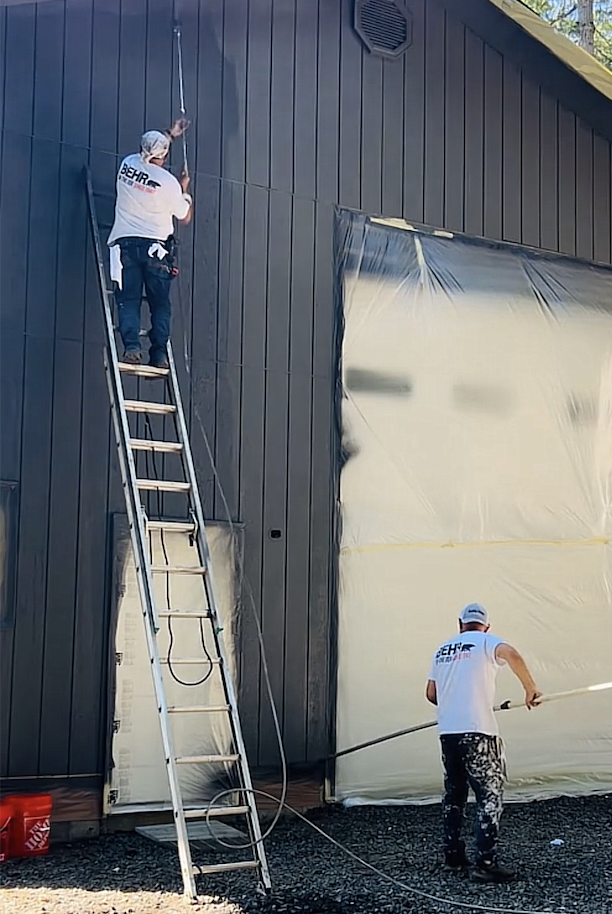Two workers painting the exterior of a dark-colored building. One worker is standing on a ladder applying paint or coating on the upper wall, the other worker is on the ground spraying paint or coating from a hose. Both are wearing white shirts, and a red bucket is on the ground.