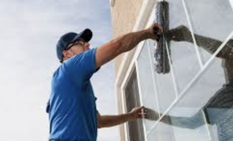 A man in a blue shirt and cap cleaning a large window with a squeegee.