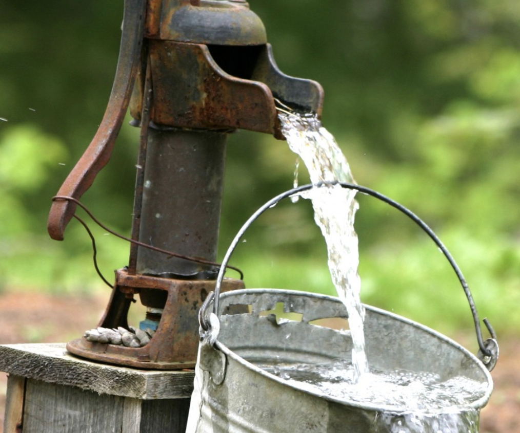 An old, rusty water pump pouring water into a metal bucket outdoors with a background of green trees and blurred foliage.