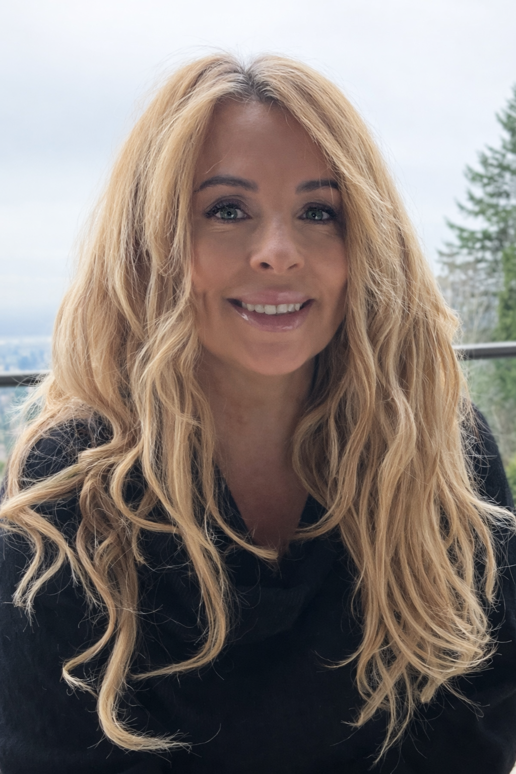Realtor Kelly Craig Graham with long, wavy blonde hair, blue eyes, and light skin, smiling, sitting on a balcony with a scenic view of trees and a cloudy sky in the background.