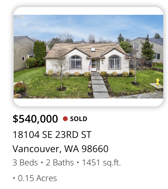 A single-story house with a beige roof and white exterior, two large arched windows in the front, a concrete walkway leading to the front door, and a landscaped front yard with shrubs and trees. Nearby houses and trees are visible in the background.