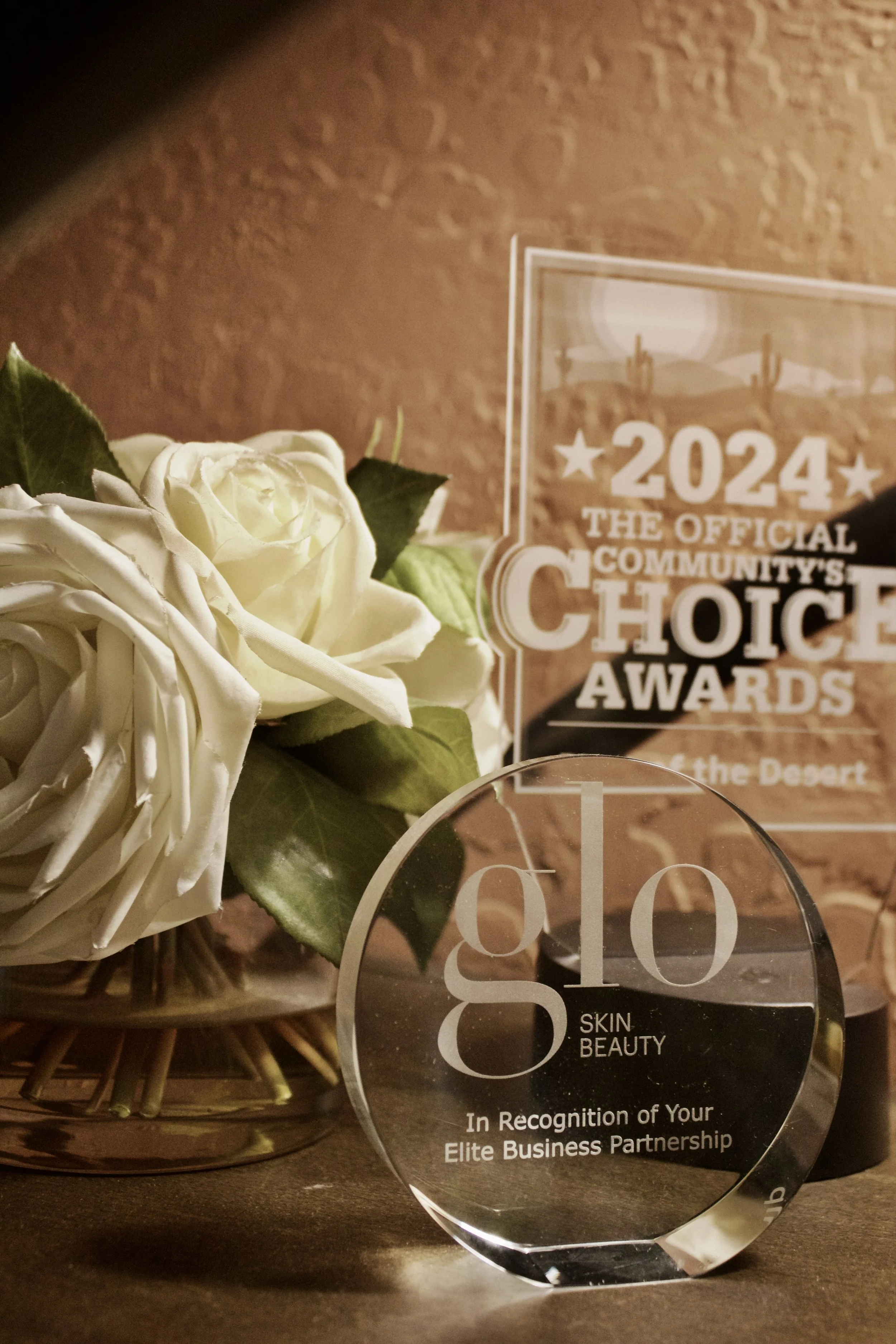 A clear crystal award with the text 'Glo Skin Beauty' and 'In Recognition of Your Elite Business Partnership.' Behind it, a bouquet of white roses with green leaves, and a glass plaque with the text '2024 The Official Community's Choice Awards' and '