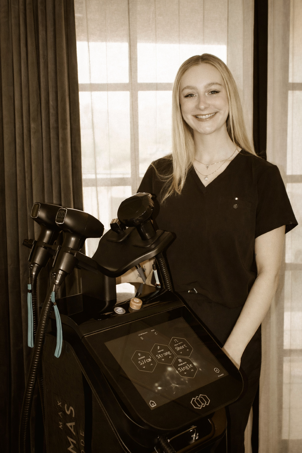 A smiling woman in medical scrubs standing next to a modern medical device with a digital screen and hand-held instruments, in a room with curtains and a window in the background.