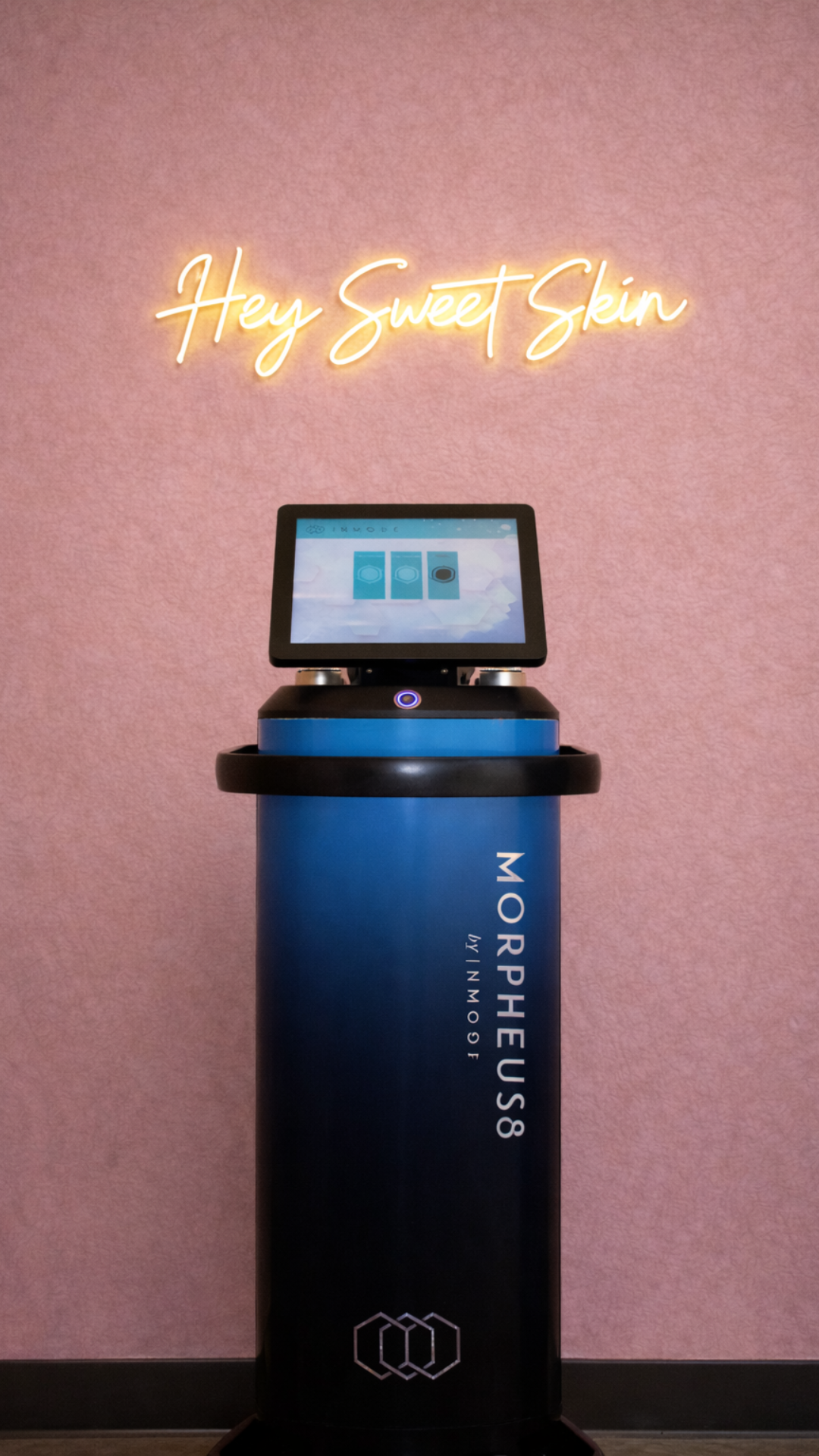 A blue and black skincare robot labeled 'MORPHEUS8 by INMODE' stands against a pink wall, with a digital screen on top displaying options, and a neon sign above reading 'Hey Sweet Skin'.