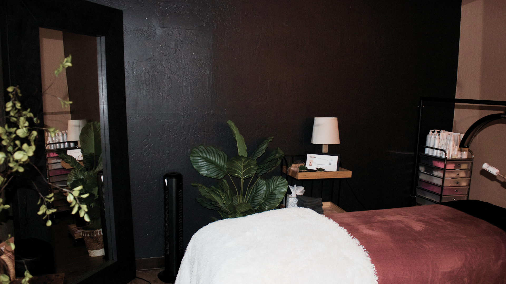 A massage room with a massage table covered in a white towel, a burgundy sheet, and green plants against a black textured wall. There is a small side table with a white lamp and beauty products.