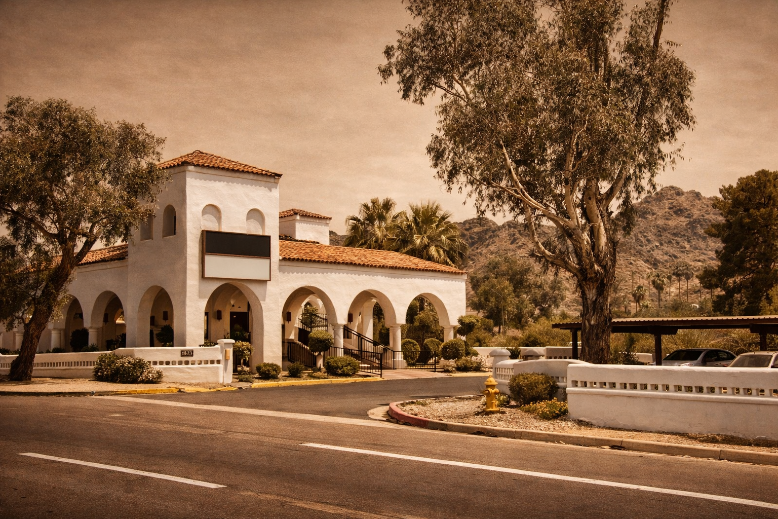 A white building with a red-tiled roof, arched doorways, and a covered porch, set against a backdrop of mountains and trees, with a street in front and a parking area to the right.
