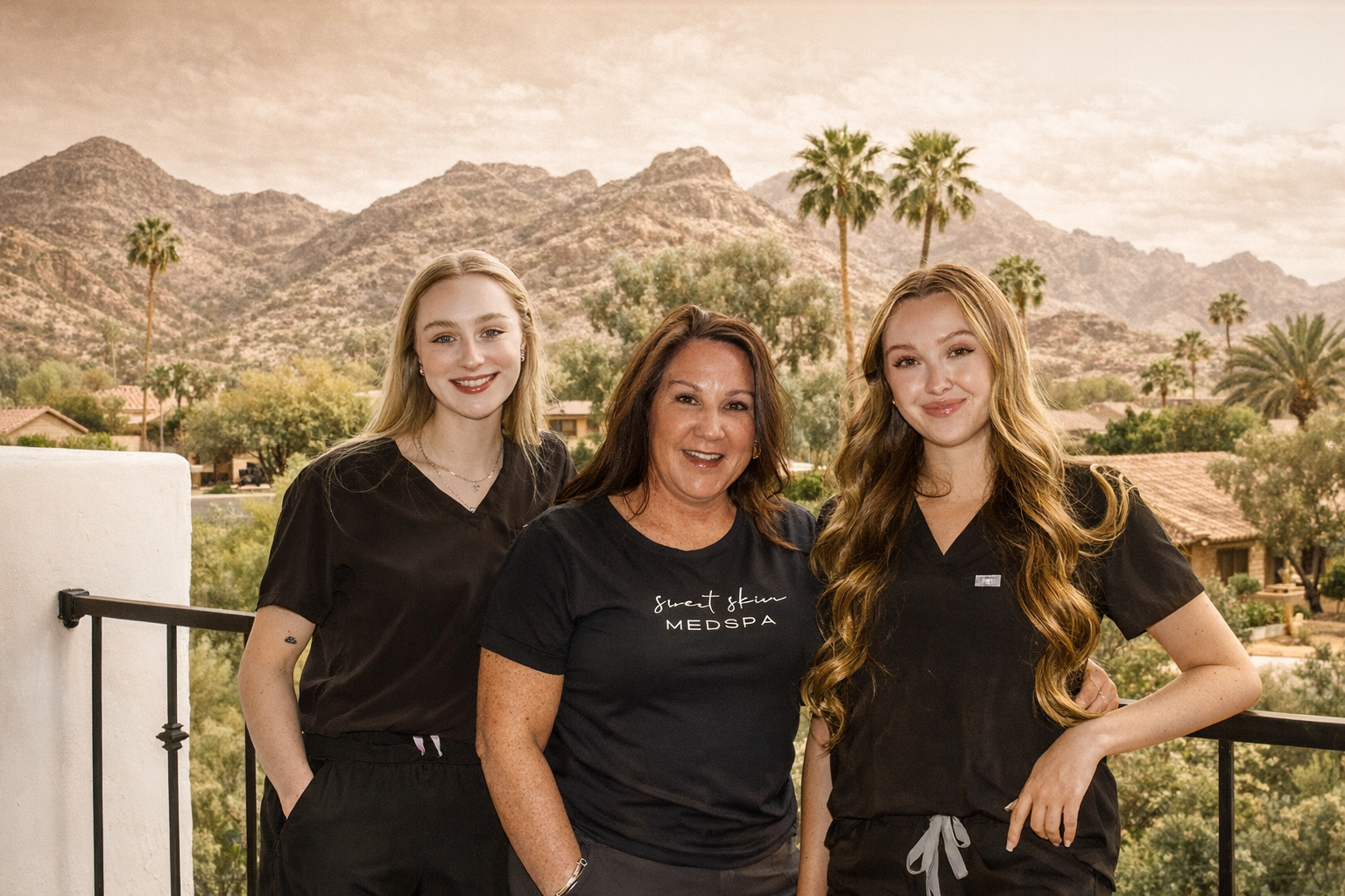 Three women standing on a balcony with a mountainous landscape and palm trees in the background. They are smiling and wearing black medical scrubs.