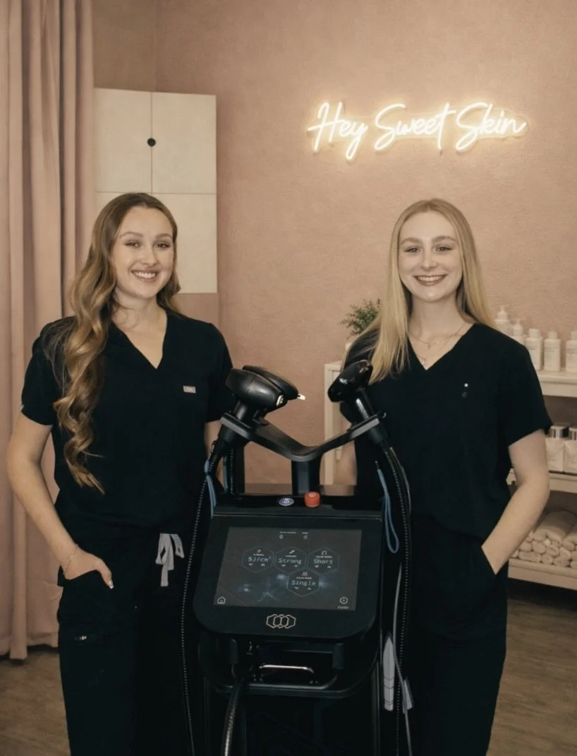 Two smiling women in black scrubs standing in a spa or skincare clinic, with a piece of equipment between them and a neon sign on the wall that says "Hey Sweet Skin."