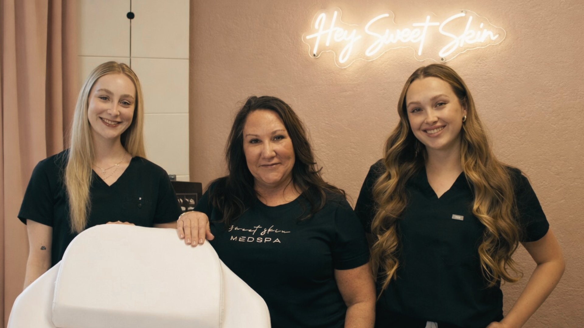 Three women in black uniforms standing together inside a spa, with a neon sign reading 'Hey Sweet Skin' on the wall behind them.