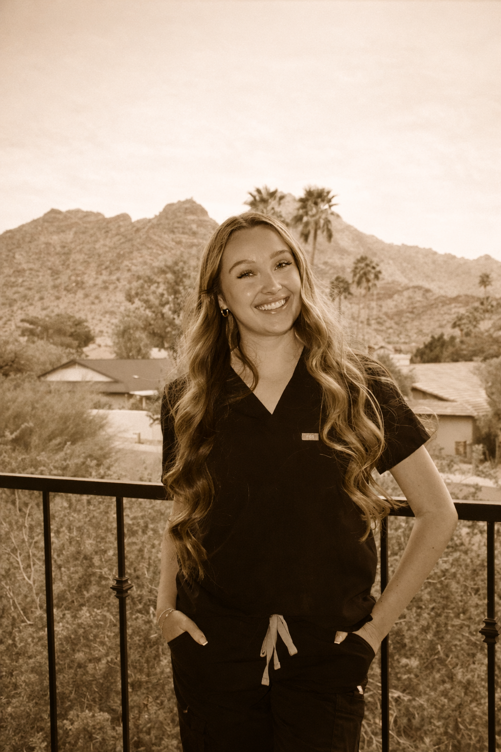 A young woman with long wavy hair standing on a balcony with mountains and palm trees in the background, smiling and wearing dark scrubs.