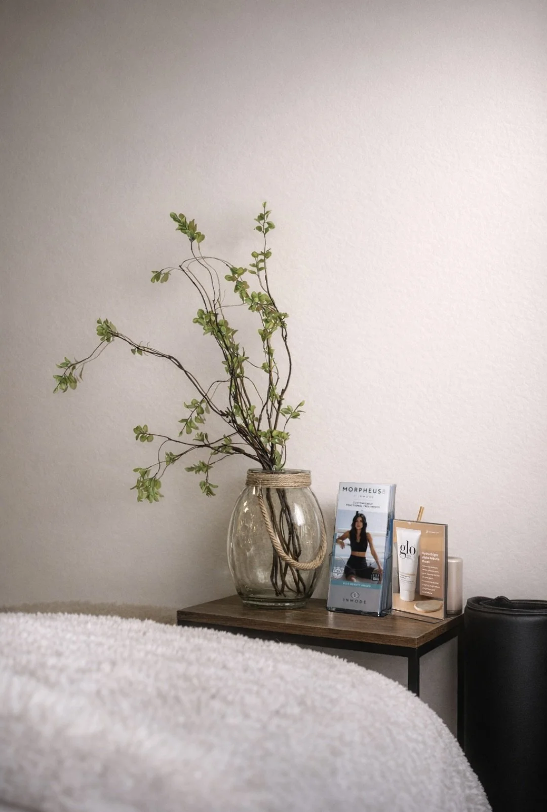 A glass vase with green branches on a wooden side table, with skincare and promotional cards beside it, against a plain white wall.