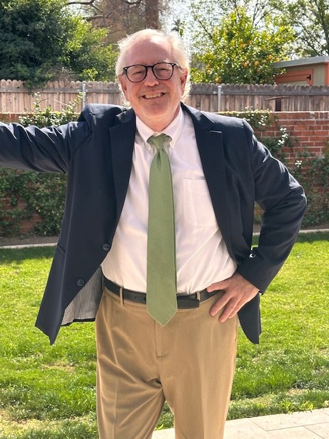 A smiling man with glasses wearing a suit jacket, white shirt, green tie, and beige pants standing outdoors in a backyard with a brick wall, trees, and bushes.