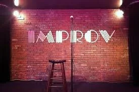 Stage with brick wall background and illuminated 'IMPROV' sign, microphone stand, and stool in front of the wall.
