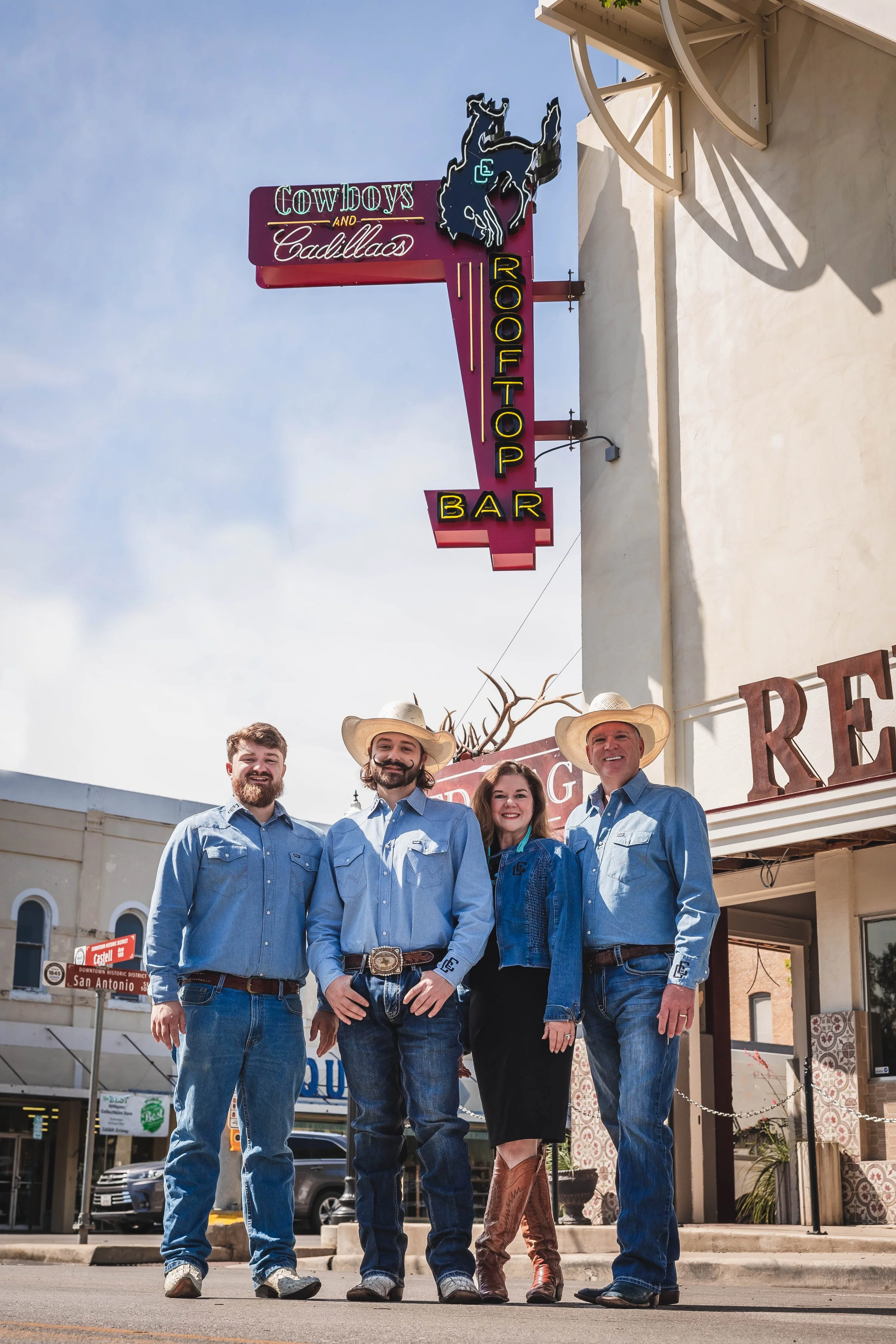 Four people dressed in western attire standing in front of a neon sign that says 'Cowboys and Cadillacs Rooftop Bar' with a cowboy hat logo. They are posing and smiling for the photo.