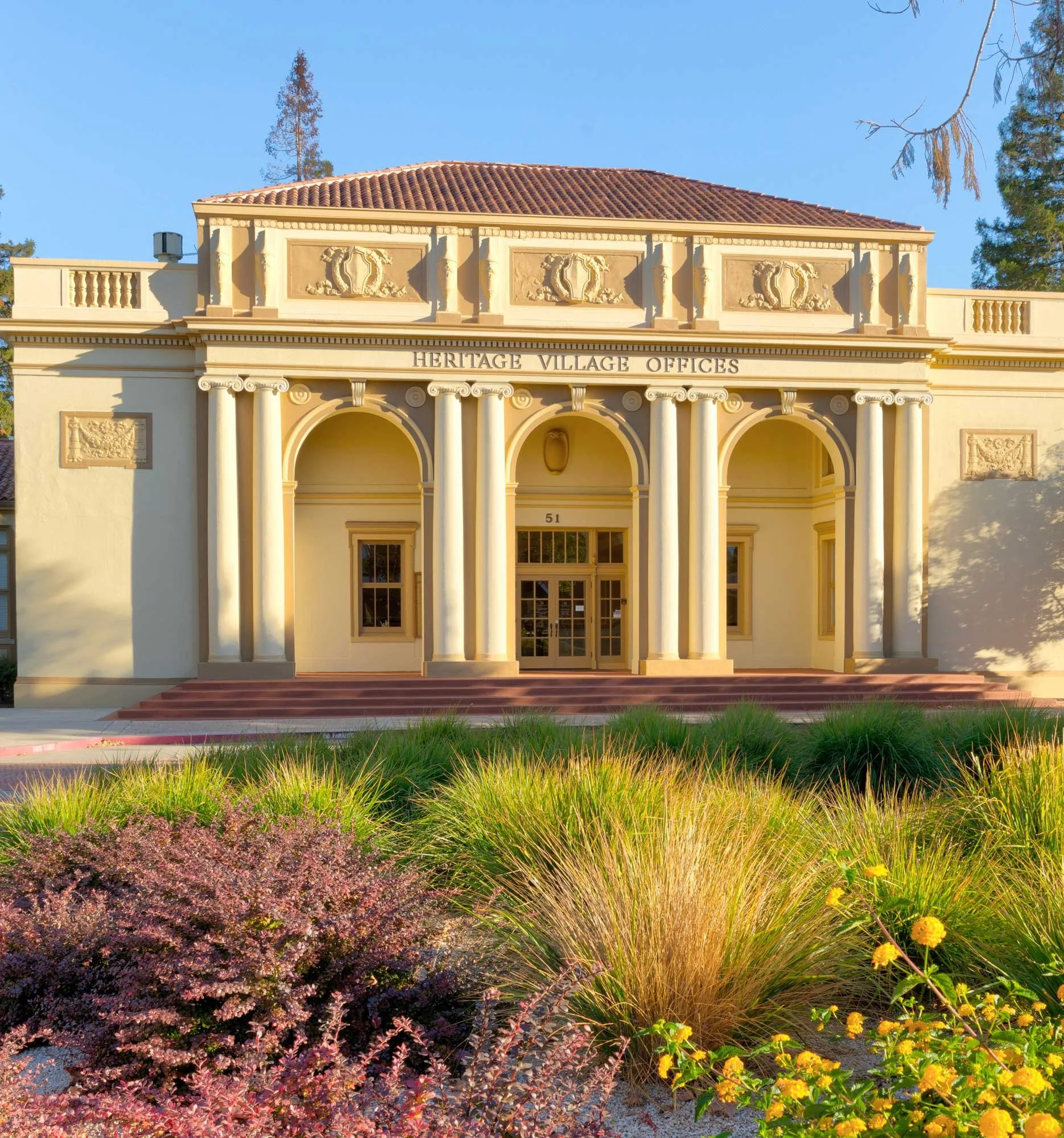 Front view of the Heritage Village Offices building with classical architecture, columns, and decorative details, surrounded by landscaped plants and bushes.