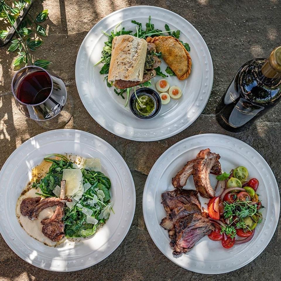 Three plates of food on a stone surface, with a glass of red wine and bottle of wine nearby. The top plate has a sandwich with greens, a fried item, a soft-boiled egg sliced in half, and a small container of sauce. The bottom-left plate contains a salad with greens, cheese shavings, and a piece of roasted meat. The bottom-right plate has grilled ribs with a side of tomato and cucumber salad.