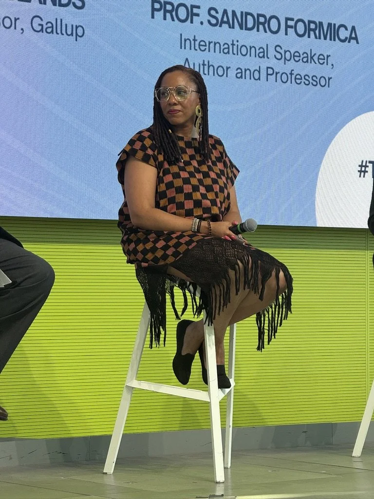 A woman sitting on a white high stool on a stage, holding a microphone, with a large blue presentation screen behind her. She is wearing glasses, earrings, a checkered dress with fringes, and black shoes with black socks.
