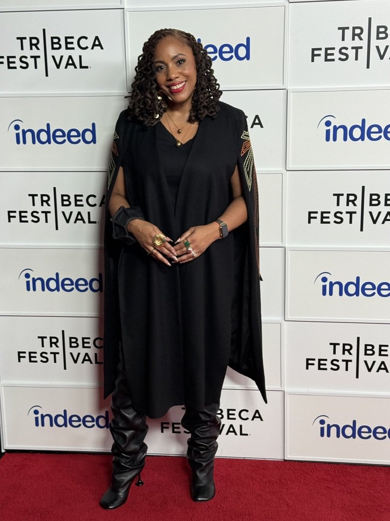 Woman standing on a red carpet at Tribeca Festival, smiling, wearing black clothing, layered necklaces, and gold rings, with a backdrop displaying Tribeca Festival and Indeed logos.