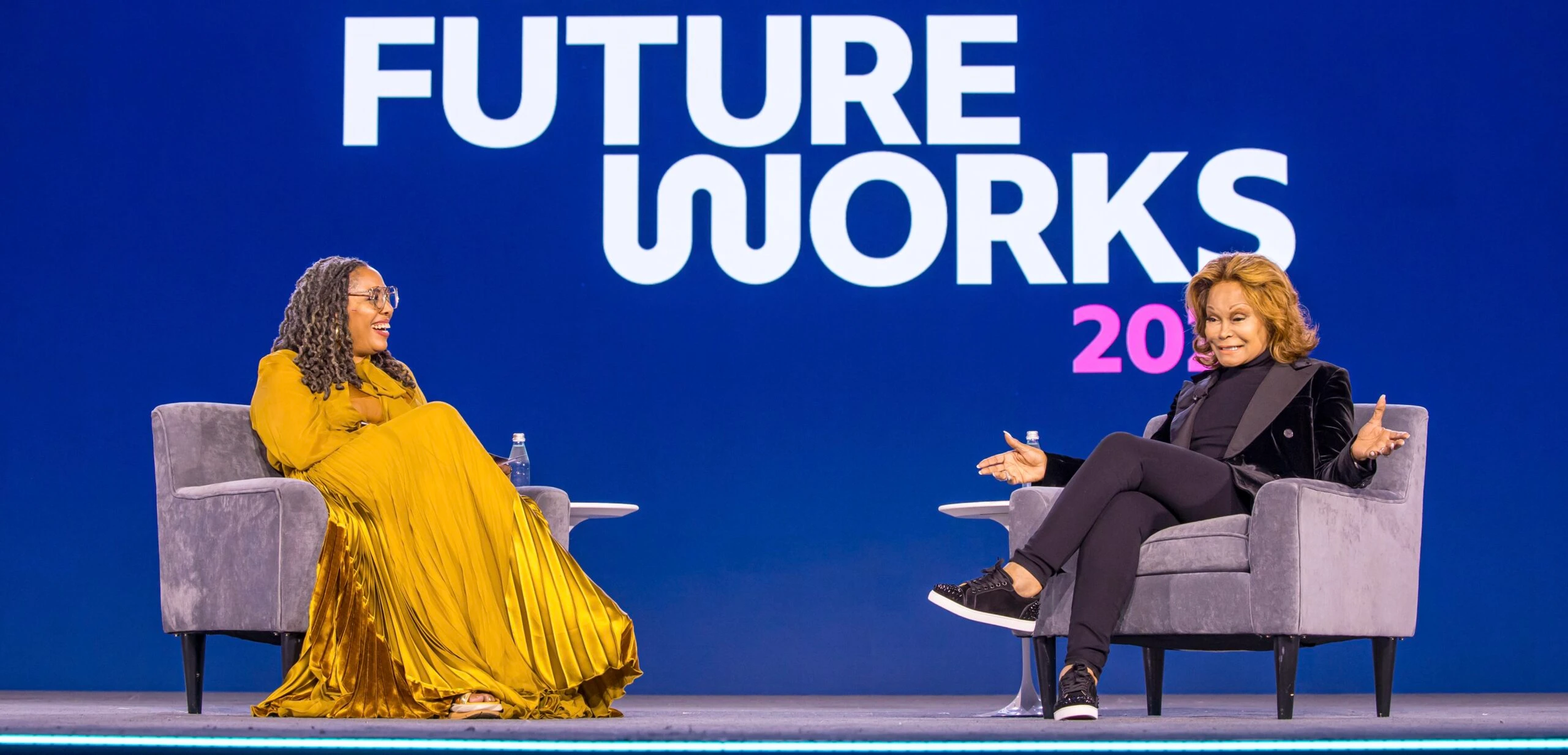 Two women in conversation on stage, sitting in gray armchairs with small tables holding water bottles, against a backdrop with 'FUTURE WORKS 202' in large text.