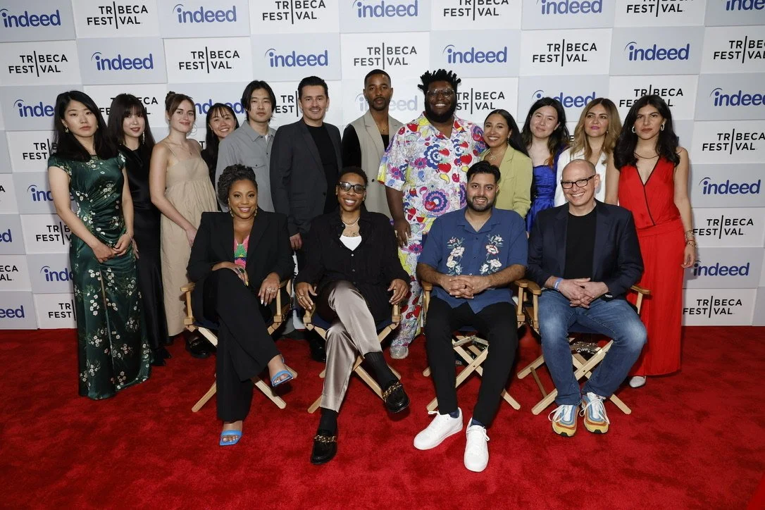 Group of diverse people at Tribeca Festival, posing for a photo in front of a backdrop with logos of Tribeca Festival, Indeed, and other sponsors, on a red carpet.