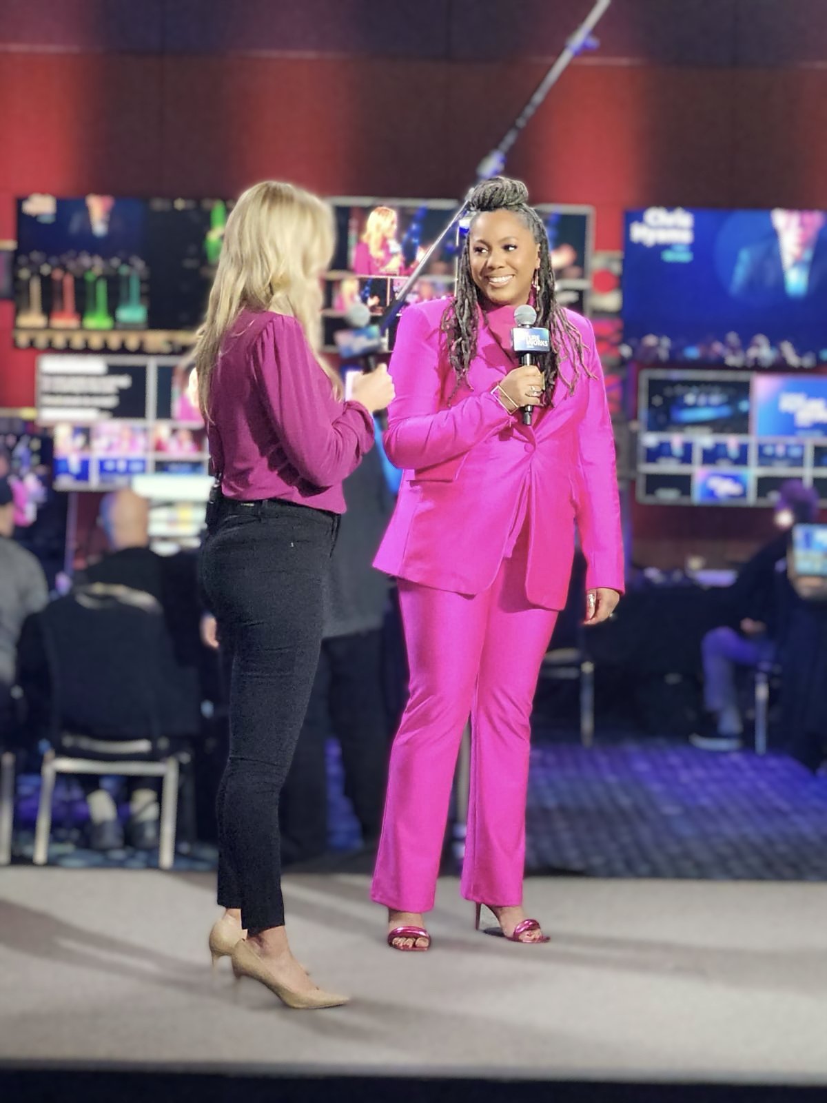 Two women on stage, one holding a microphone, engaged in conversation. The woman on the right is wearing a bright pink suit, and the woman on the left is wearing a purple top and black pants. Large screens and cameras are in the background, indicatin