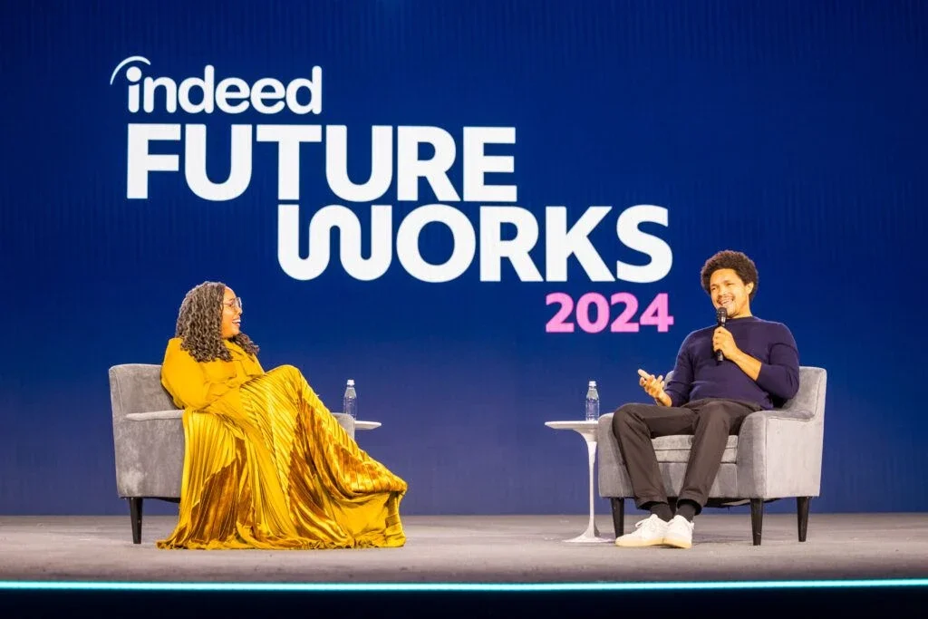 A woman and a man are sitting on stage chairs, engaging in a discussion at the 'Indeed Future Works 2024' event with a large blue backdrop behind them.