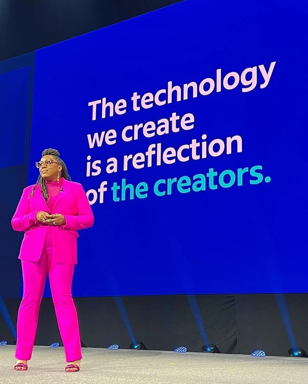 A woman in a bright pink suit speaking at a conference with a large blue screen behind her displaying a quote about technology and creators.
