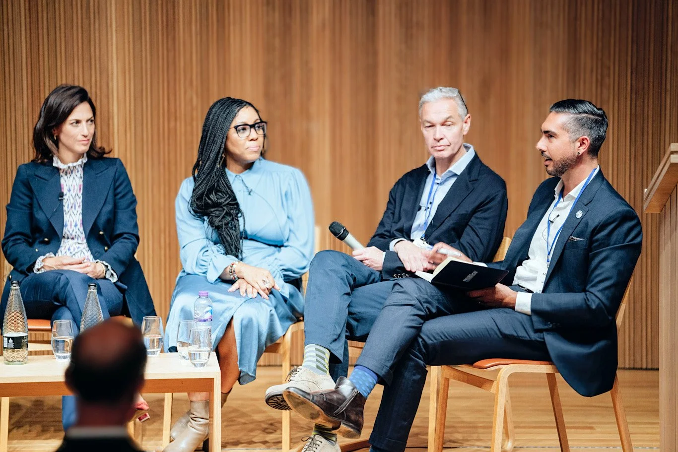 Panel discussion with four people sitting on stage in front of a wood-paneled wall, with microphones and water bottles on tables in front of them.