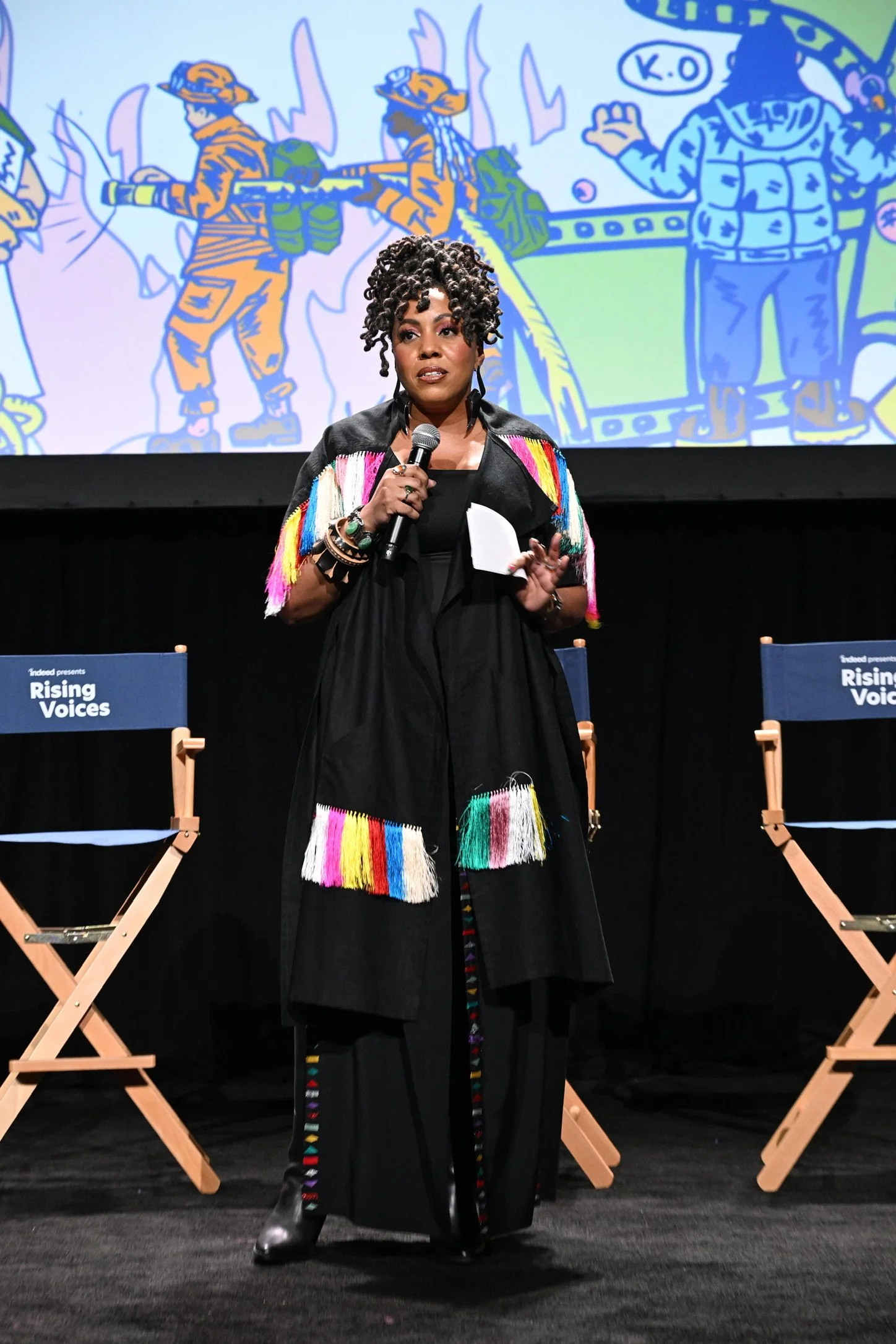 A woman with short curly hair speaking into a microphone on stage, wearing a black outfit with colorful tassels. Behind her is a large illustration of soldiers and a cartoon cowboy with a speech bubble. Two chairs with 'Rising Voices' signs are on ei