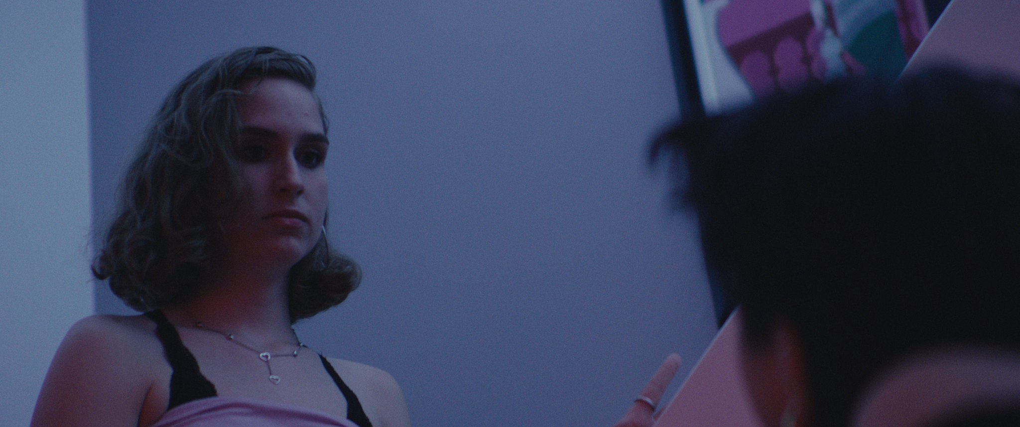 A woman with shoulder-length curly hair looking at a person, with purple and pink lighting in the background.