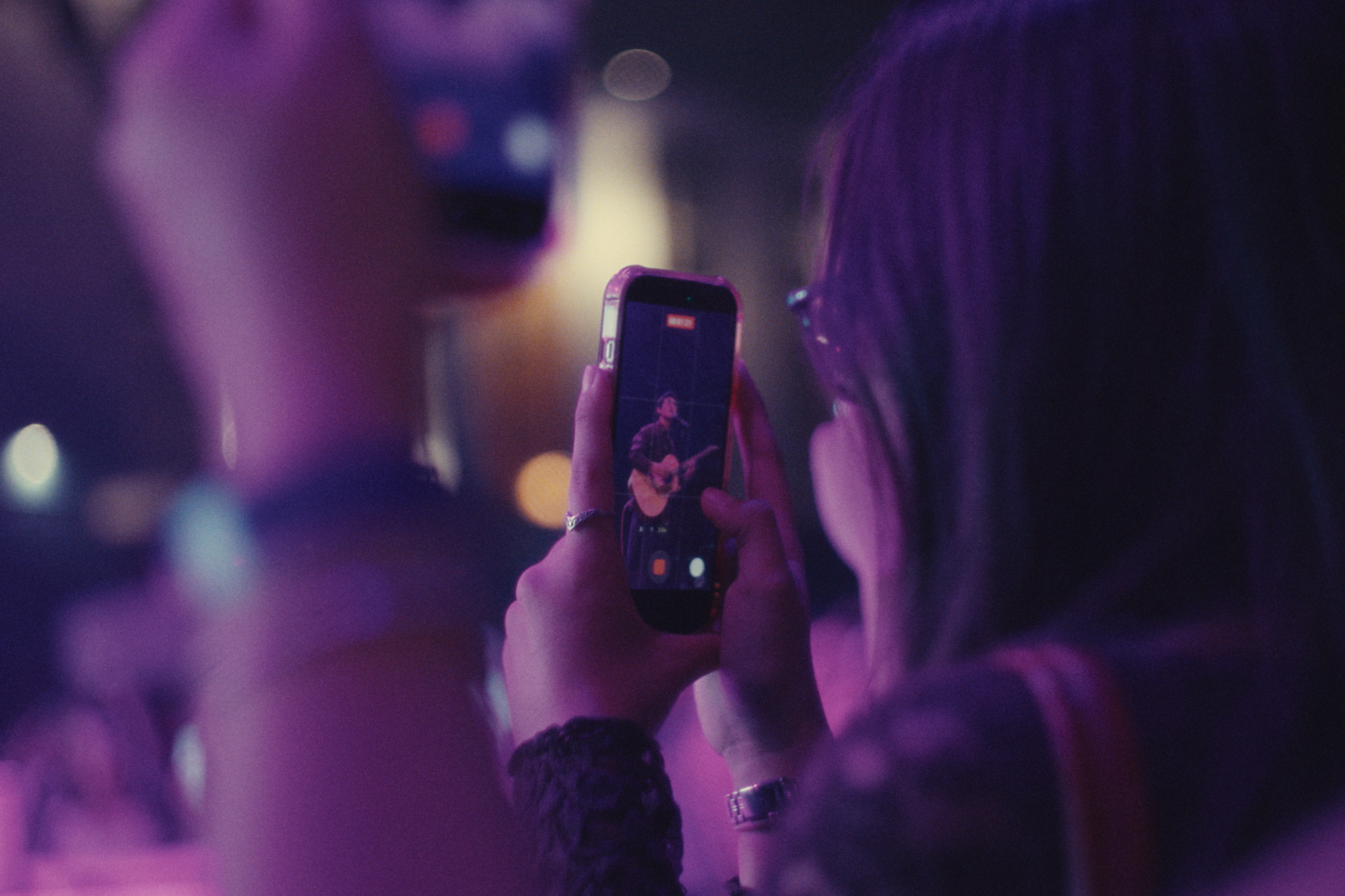 A woman taking a photo or recording a video of a musician playing guitar on stage with a smartphone during a live music event, illuminated by purple and pink stage lighting.