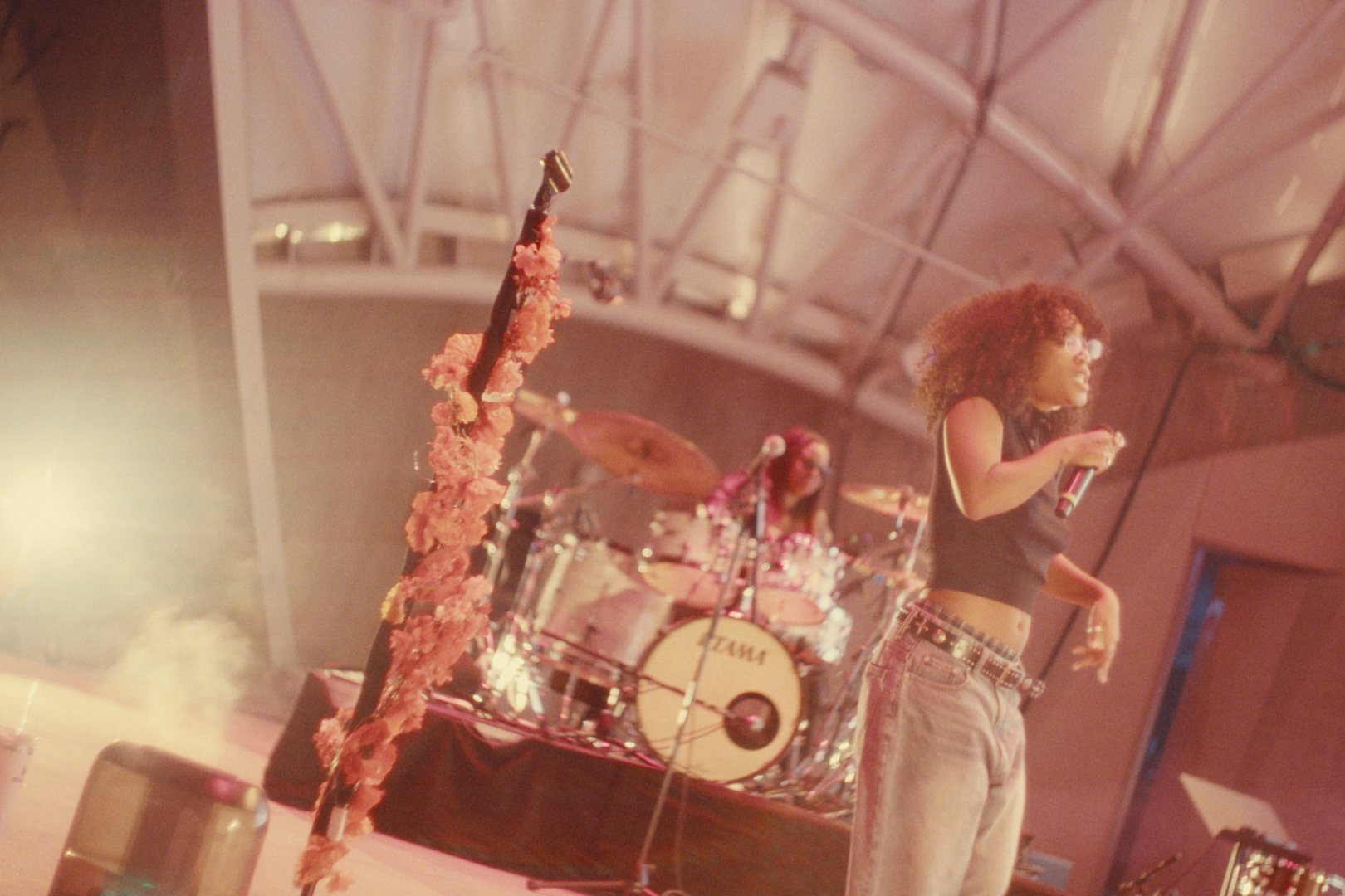 A woman with curly hair, wearing glasses, a black crop top, and baggy jeans, performs with a microphone on stage. Behind her, a drummer with long hair plays a drum set decorated with pink foliage.