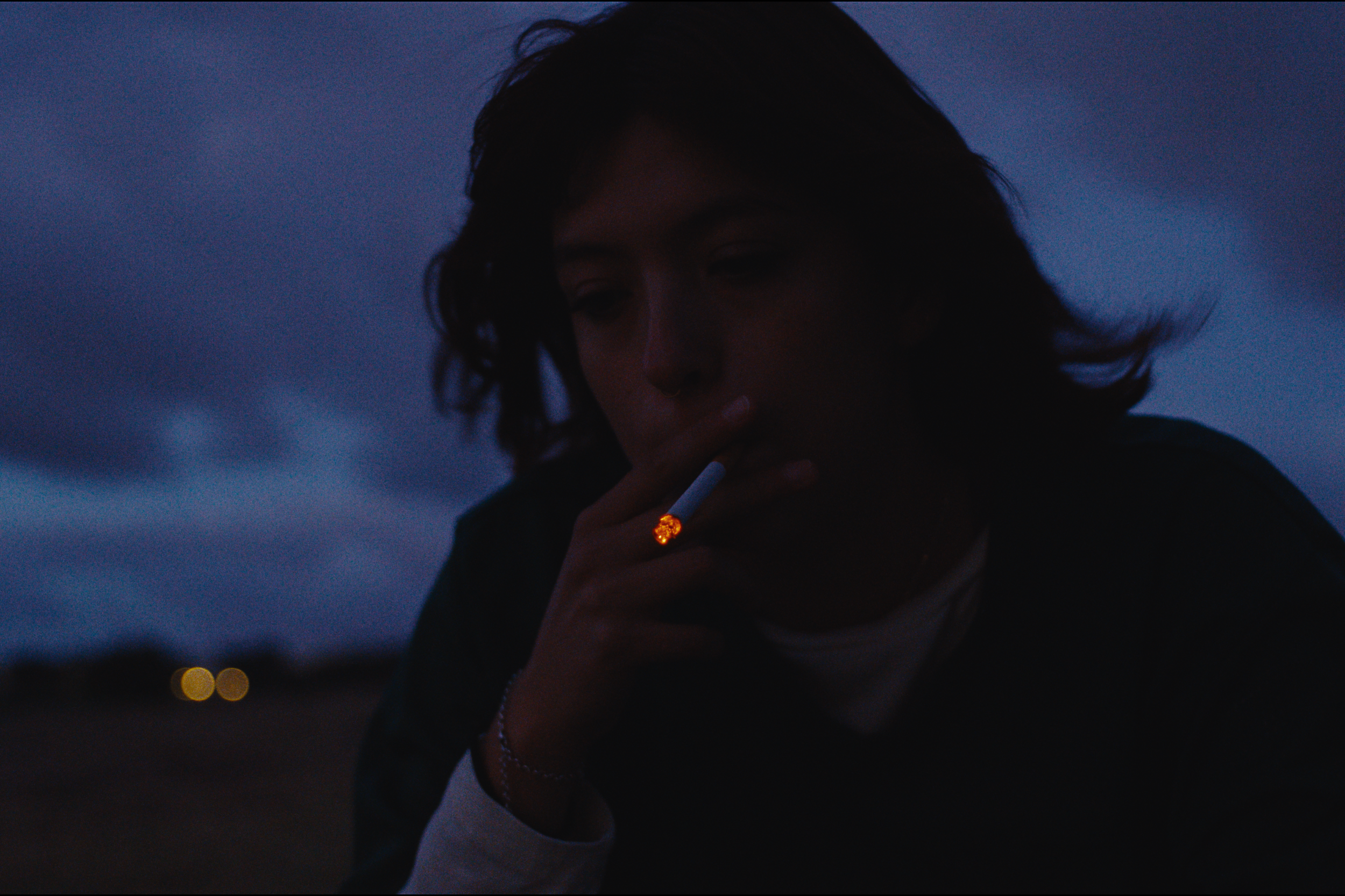 A person with dark hair smoking a cigarette at dusk, with a dark blue sky and blurred city lights in the background.