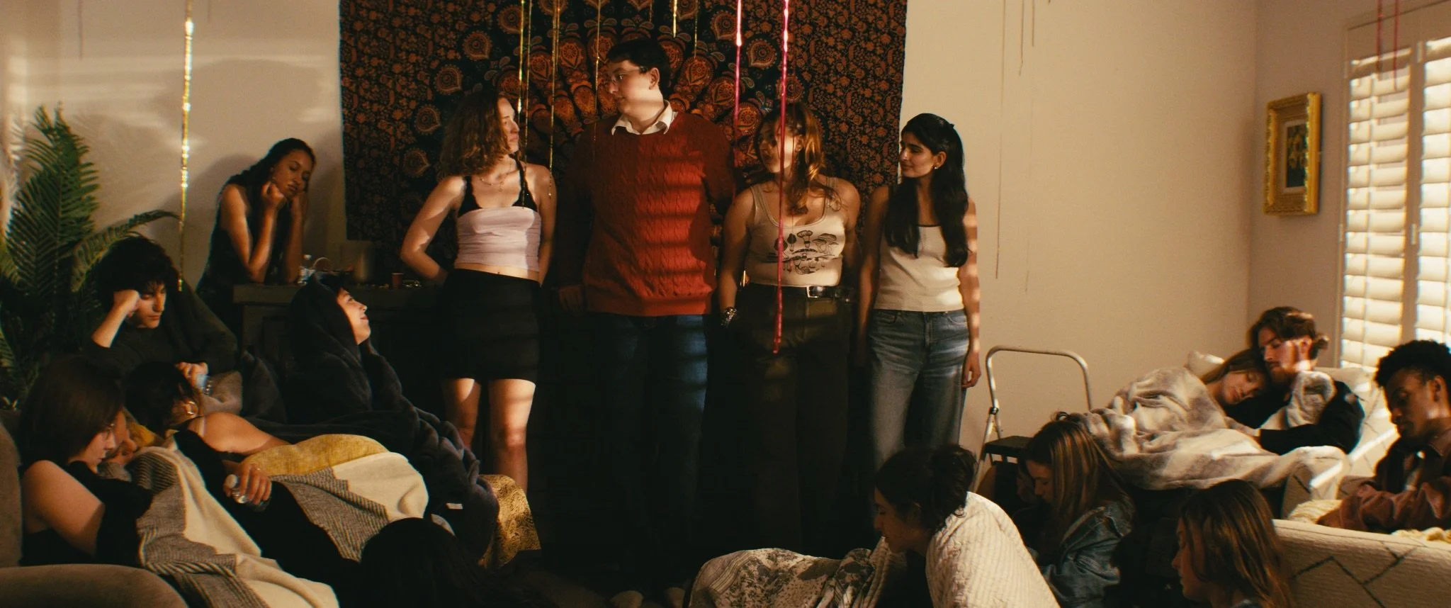 Group of young people gathered in a cozy living room, some sitting on couches and others standing, with some asleep and others chatting, during a casual party or gathering.