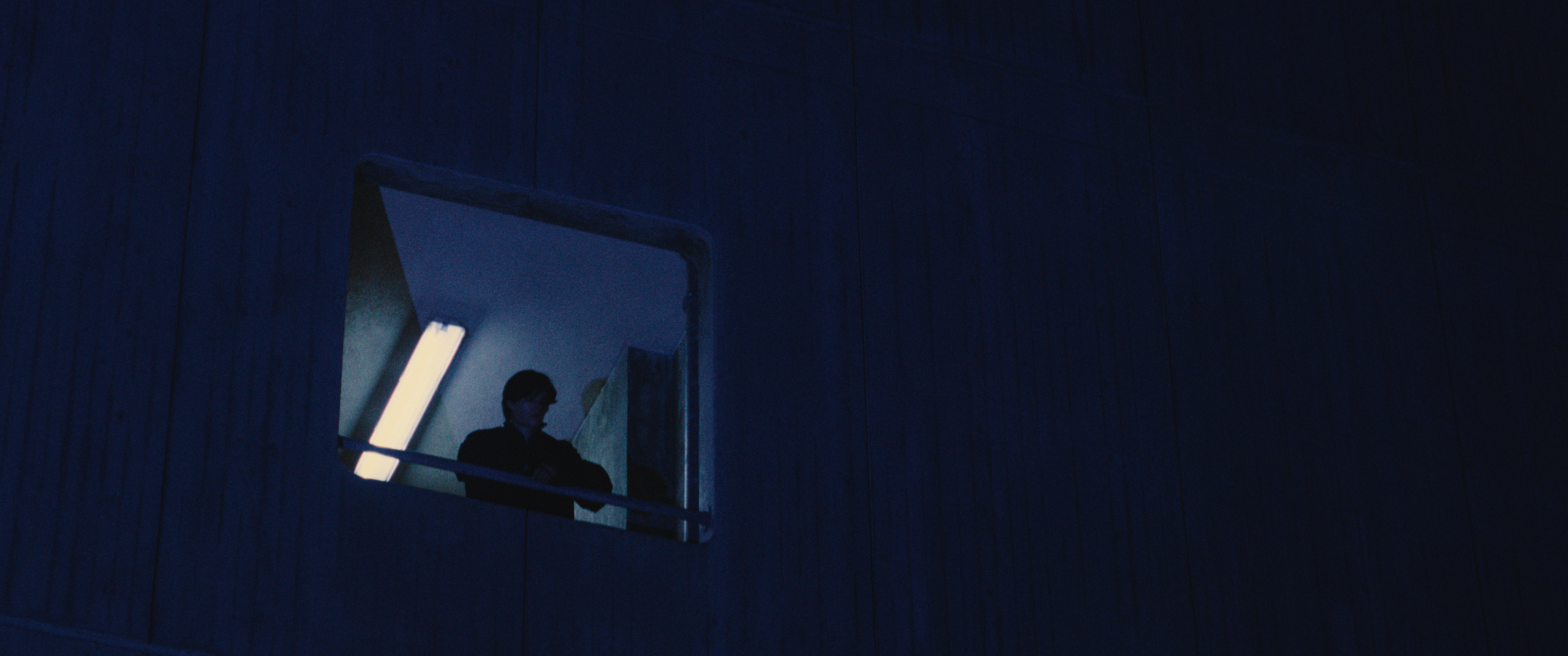 A person sitting in a small illuminated room or booth, seen through a window set in a dark wooden wall, with blue lighting creating a moody atmosphere.