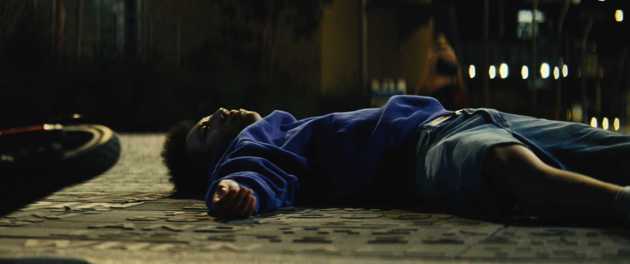 A young person lying on the sidewalk at night with a bicycle tire nearby, wearing a blue hoodie and shorts, surrounded by dark surroundings and city lights.