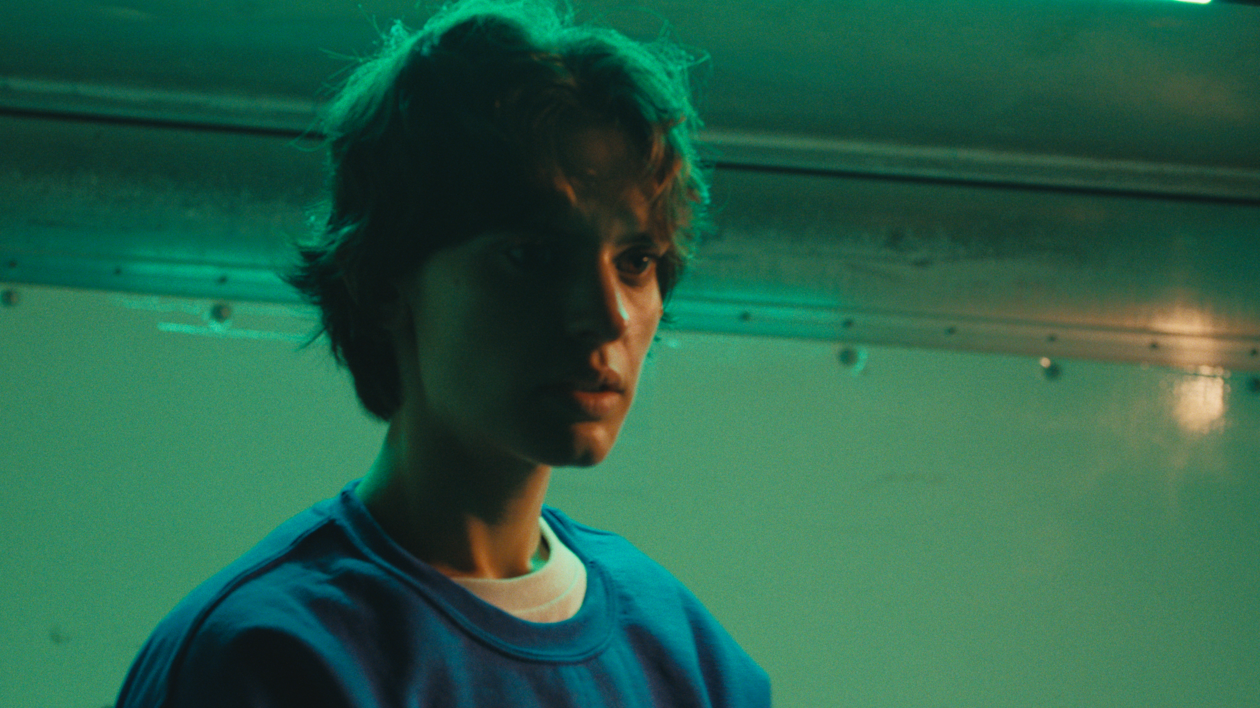 A young person with short, tousled hair looking at the camera against a greenish background with metallic ceiling panels.
