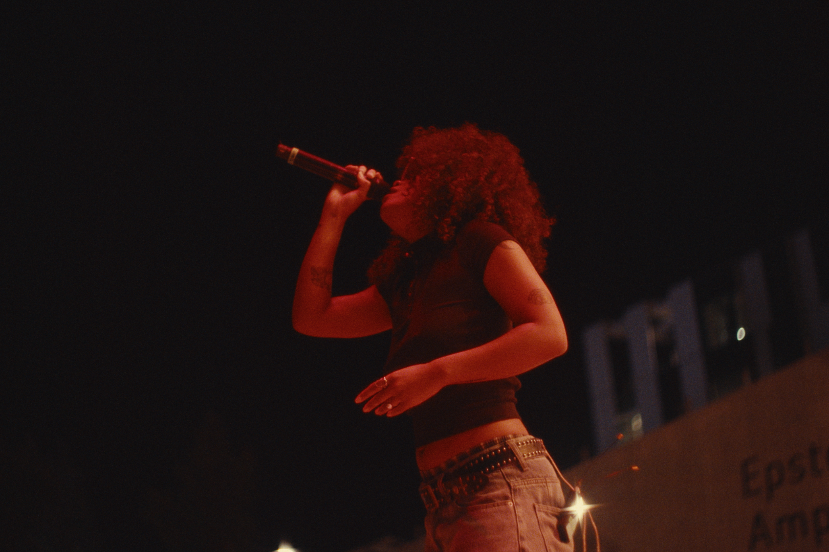 A woman with curly hair holding a microphone to her mouth, singing or speaking at night, with buildings visible in the background.