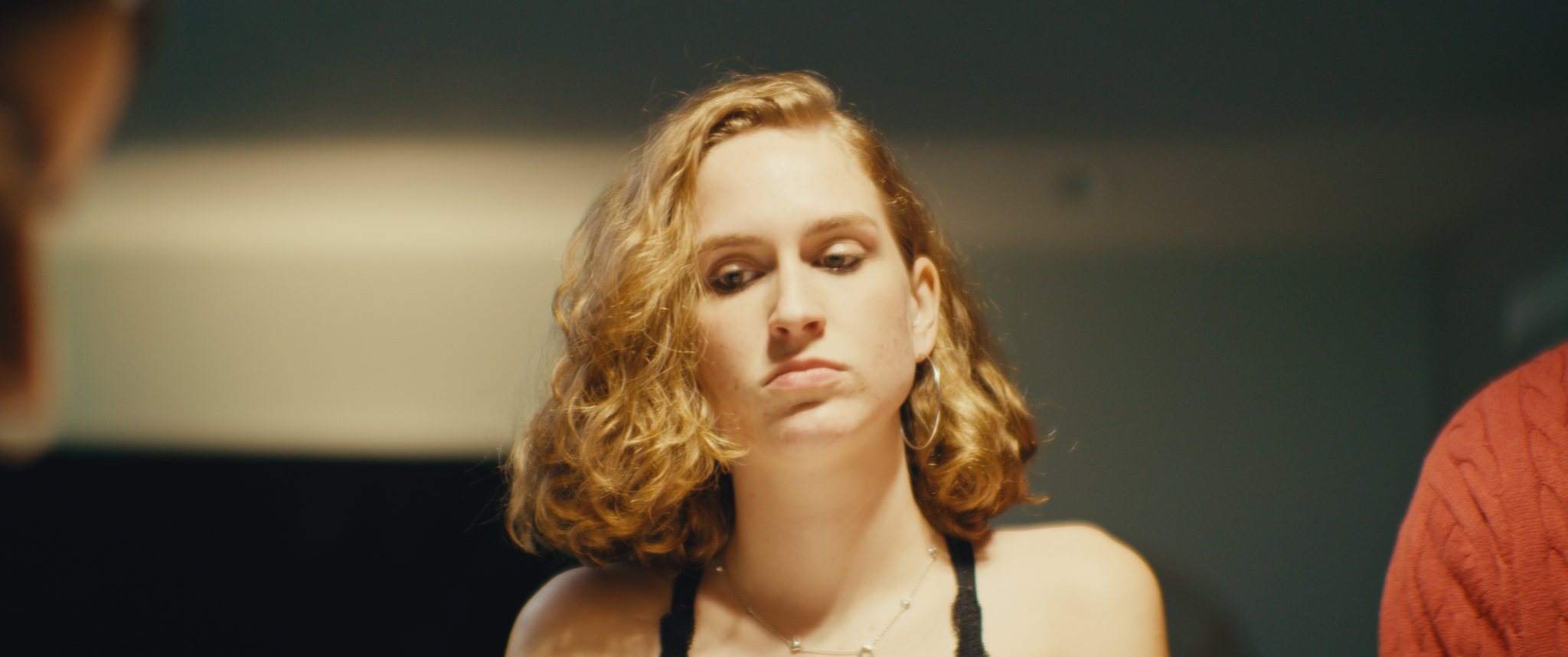 A young woman with curly blonde hair looking down with a serious expression, wearing a black strappy top and jewelry, in a dimly lit indoor setting.