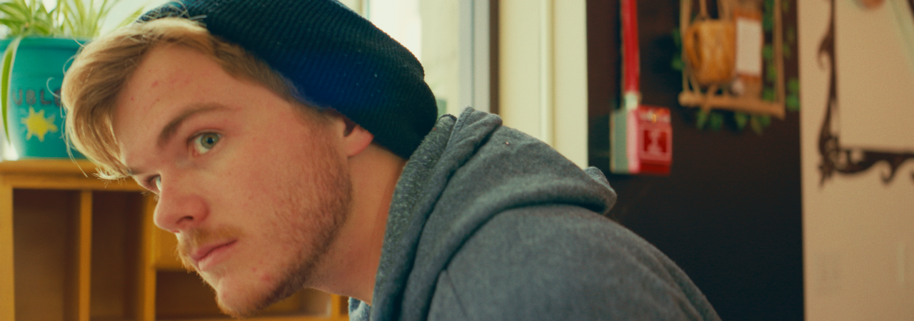 A close-up of a young man wearing a black beanie and a gray hoodie, looking sideways in an indoor setting with a blurred background of shelves and plants.